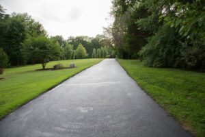 Freshly paved asphalt pathway running through a landscaped park setting, bordered by green grass, trees, and shrubs under an overcast sky.